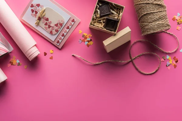 Top view of valentines decoration, wrapping paper, twine, gift box with chocolate, greeting card on pink background — Stock Photo