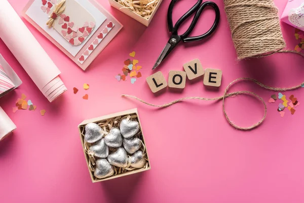 Vista superior de la decoración de San Valentín, tijeras, papel de envolver, cordel, caja de regalo con corazones de plata, tarjeta de felicitación y letras de amor en cubos de madera sobre fondo rosa - foto de stock
