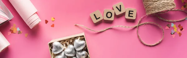 Vue du dessus du papier d'emballage, ficelle, boîte cadeau avec des cœurs argentés et lettrage d'amour sur des cubes en bois sur fond rose, prise de vue panoramique — Photo de stock