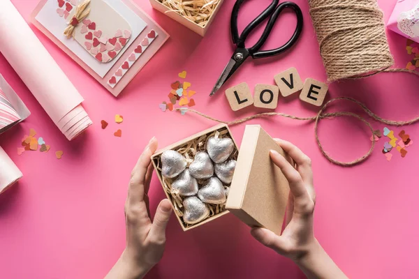 Partial view of woman holding gift box with silver hearts near valentines handiwork supplies on pink background — Stock Photo