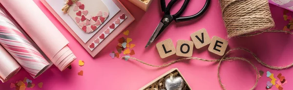 Top view of valentines decoration, scissors, wrapping paper, twine, gift, greeting card and love lettering on wooden cubes on pink background, panoramic shot — Stock Photo