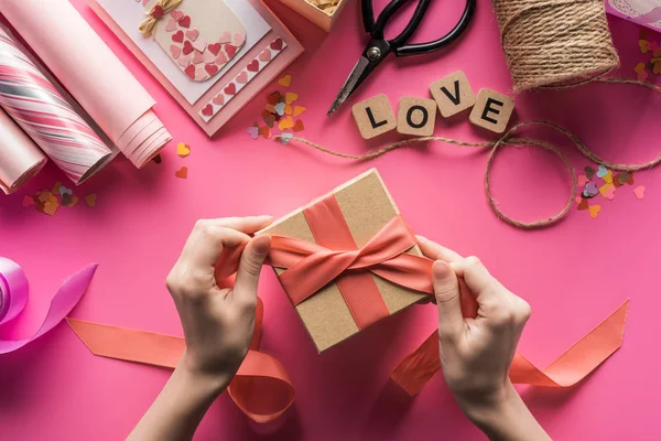 Vue partielle de la femme décorant boîte cadeau avec ruban près de fournitures de bricolage valentines sur fond rose — Photo de stock
