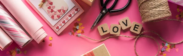 Top view of valentines decoration, scissors, wrapping paper, twine, gift, greeting card and love lettering on wooden cubes on pink background, panoramic shot — Stock Photo