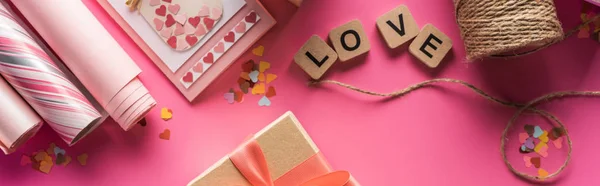 Top view of valentines decoration, wrapping paper, twine, gift, greeting card and love lettering on wooden cubes on pink background, panoramic shot — Stock Photo