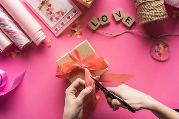 Cropped view of woman cutting ribbon while making valentines gift on pink background — Stock Photo