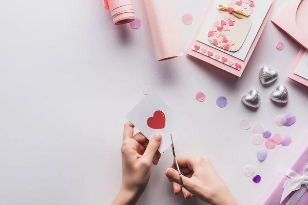 Vista recortada de la mujer cortando el corazón con tijeras cerca de la decoración de San Valentín, regalos, corazones y papel de envolver sobre fondo blanco - foto de stock