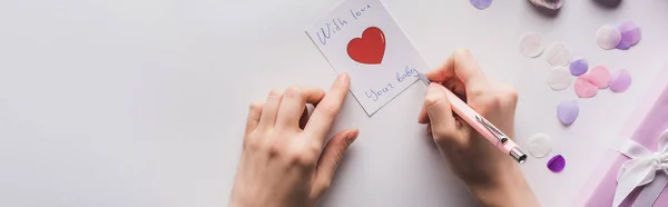 Vue recadrée d'une femme écrivant sur une carte près d'un cadeau de Saint-Valentin et confettis sur fond blanc, vue panoramique — Photo de stock