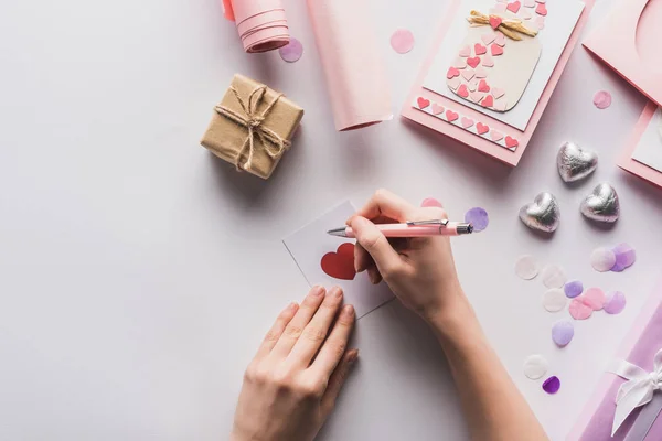 Vista recortada de la mujer escribiendo en la tarjeta cerca de la decoración de San Valentín, regalos, corazones y papel de embalaje sobre fondo blanco - foto de stock