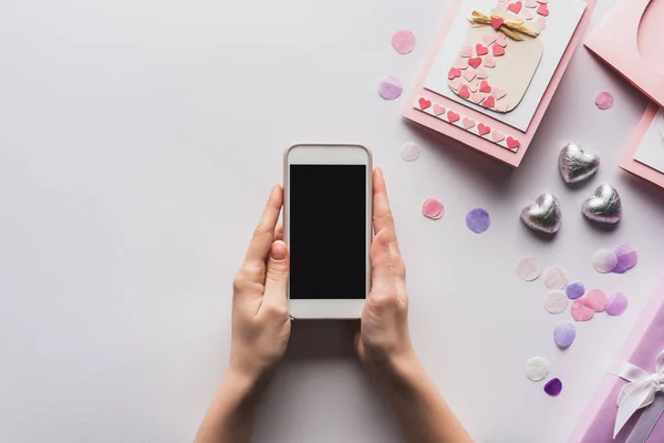 Vista parcial de la mujer sosteniendo teléfono inteligente cerca de regalos de San Valentín y decoración sobre fondo blanco - foto de stock