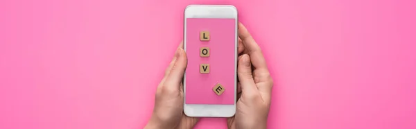 Partial view of woman holding smartphone with image of cubes with love lettering on pink background, panoramic shot — Stock Photo