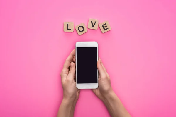 Teilansicht einer Frau, die ihr Smartphone in der Nähe von Würfeln mit Liebesschrift auf rosa Hintergrund hält — Stockfoto