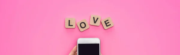 Vista parcial de la mujer sosteniendo teléfono inteligente cerca de cubos con letras de amor sobre fondo rosa, plano panorámico - foto de stock