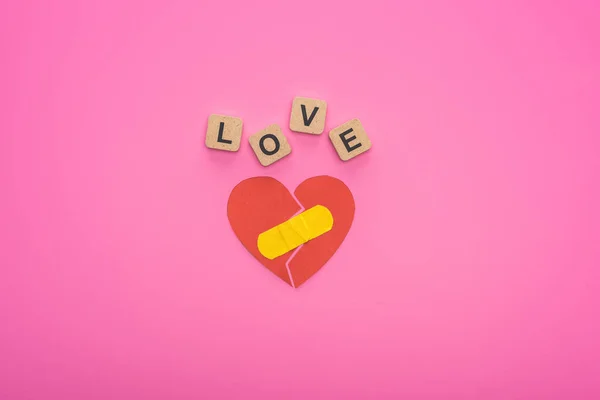 Vista dall'alto di amore lettering su cubi di legno vicino cuore spezzato con patch su sfondo rosa — Foto stock