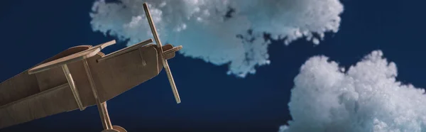 Avión de juguete de madera volando entre nubes esponjosas blancas hechas de algodón aislado en azul oscuro, plano panorámico - foto de stock