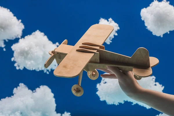 Vista recortada de la mujer sosteniendo avión de juguete de madera cerca de nubes esponjosas blancas hechas de algodón aislado en azul - foto de stock