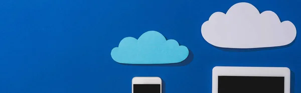 Vista superior de nubes de papel vacías cerca de smartphone y tableta digital con pantalla en blanco aislada en azul, plano panorámico - foto de stock