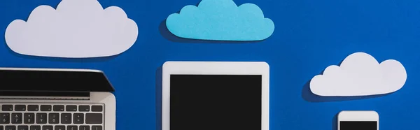 Vista superior de nubes de papel vacías cerca del ordenador portátil, smartphone y tableta digital con pantalla en blanco aislada en azul, plano panorámico - foto de stock