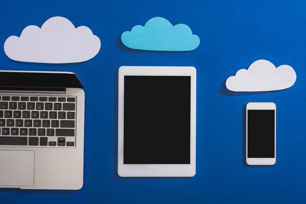 Vista superior de nubes de papel vacías cerca del portátil, smartphone y tableta digital con pantalla en blanco aislada en azul - foto de stock