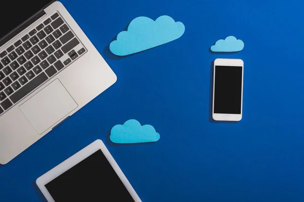 Vista superior de nuvens de papel vazias perto de laptop, smartphone e tablet digital com tela em branco isolado em azul — Fotografia de Stock