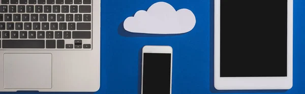 Top view of empty white paper cloud near laptop, smartphone and digital tablet with blank screen isolated on blue, panoramic shot — Stock Photo