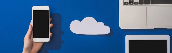 Vista recortada de la mujer sosteniendo teléfono inteligente cerca de la nube de papel blanco vacío y portátil con tableta digital en azul, plano panorámico - foto de stock