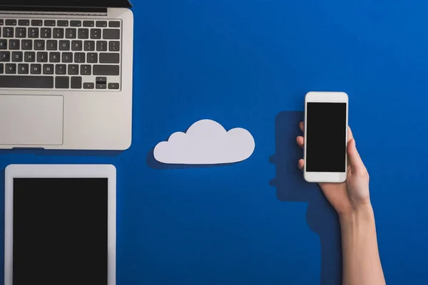 Cropped view of woman holding smartphone near empty white paper cloud and laptop with digital tablet on blue — Stock Photo