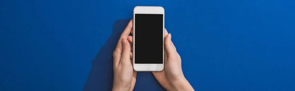 Обрезанный вид женщины, держащей смартфон на синем фоне, панорамный снимок — стоковое фото