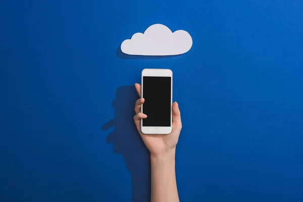 Vista recortada de la mujer sosteniendo teléfono inteligente cerca de la nube de papel blanco vacío en azul - foto de stock