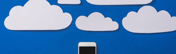 Top view of white paper cut clouds and smartphone on blue background, panoramic shot — Stock Photo
