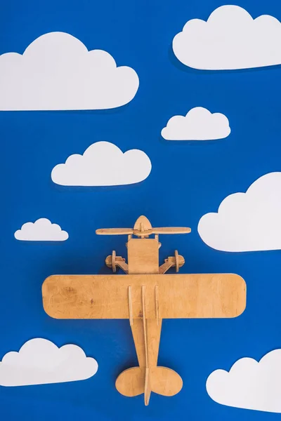 Vista superior de avión de juguete de madera en el cielo azul con papel cortado nubes blancas - foto de stock