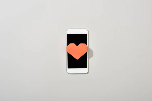 Vista superior del corazón de papel en smartphone con pantalla en blanco sobre fondo gris - foto de stock