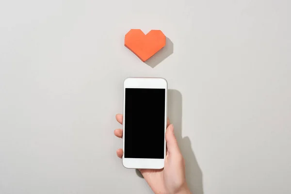 Top view of girl holding smartphone with blank screen by heart shaped paper on grey background — Stock Photo