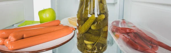 Panoramic shot of sausages, pickles and chili peppers in fridge with open door isolated on white — Stock Photo