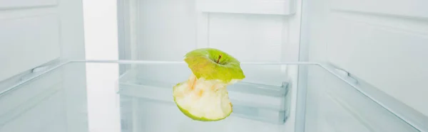 Tiro panorâmico de maçã verde roída na prateleira do refrigerador com porta aberta isolada em branco — Fotografia de Stock