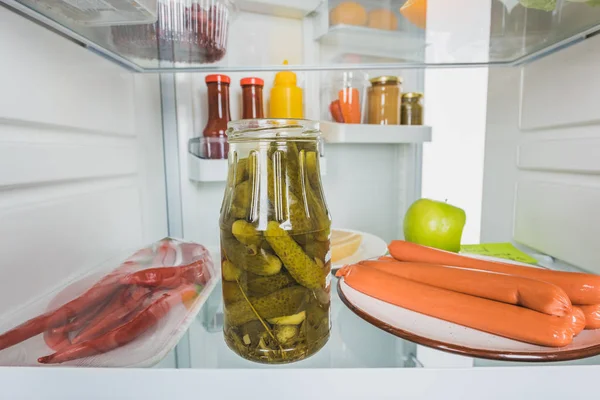 Glas mit Essiggurken, Chilischoten mit Würstchen und Soßen im Kühlschrank mit offener Tür isoliert auf weiß — Stockfoto