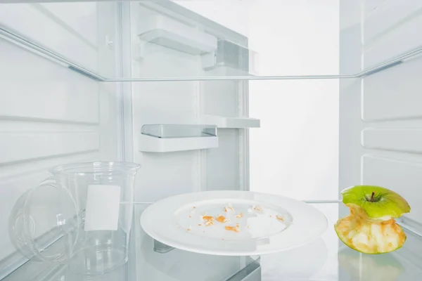 Plastic cup with bitten apple and crumbs on plate in fridge with open door isolated on white — Stock Photo