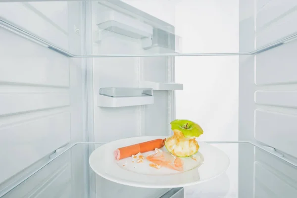 Pomme grignotée aux saucisses et miettes dans une assiette au réfrigérateur avec porte ouverte isolée sur fond blanc — Photo de stock
