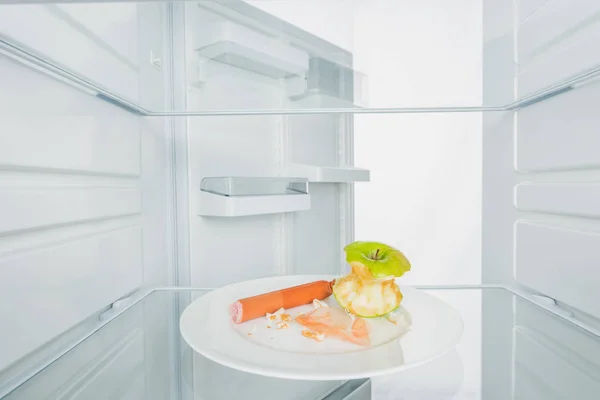 Gnawed apple with sausage and crumbs on plate in refrigerator with open door isolated on white — Stock Photo