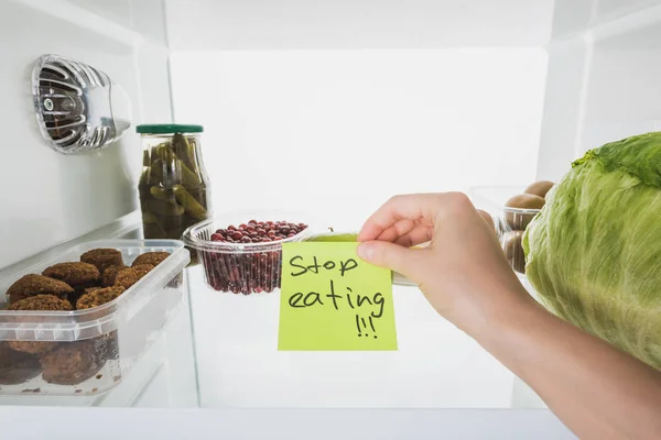 Cropped view of woman holding card with stop eating lettering with food in fridge at background, isolated on white — Stock Photo