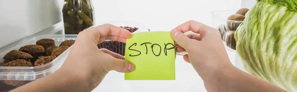 Fotografia panorâmica de mulher segurando cartão com stop lettering com alimentos na geladeira no fundo, isolado em branco — Fotografia de Stock