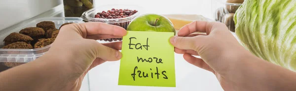 Panorâmica tiro de mulher segurando cartão com comer mais frutas lettering com alimentos na geladeira no fundo, isolado em branco — Fotografia de Stock