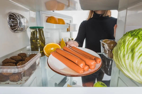 Vista cortada de mulher tomando prato com salsichas de geladeira com alimentos isolados em branco — Fotografia de Stock
