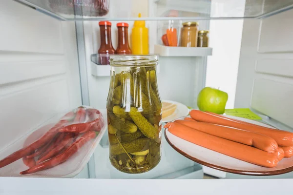 Selective focus of sausages with chili peppers and pickles in fridge with open door isolated on white — Stock Photo