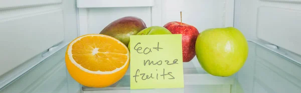 Panoramic shot of fresh fruits and cart with eat more fruits lettering on fridge shelf — Stock Photo