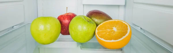 Tiro panorâmico de manga fresca com maçãs e fatia de laranja na prateleira do refrigerador — Fotografia de Stock