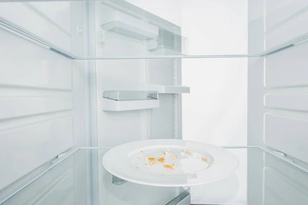 Тарелка с крошками в холодильнике с открытой дверью, изолированной на белом — стоковое фото