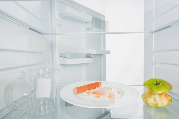 Plate with scraps, bitten apple and plastic cup in fridge with open door isolated on white — Stock Photo