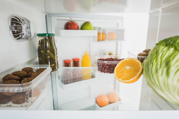 Cutlets with ripe fruits and cabbage in refrigerator with open door isolated on white — Stock Photo