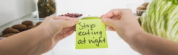 Cropped view of woman holding card with stop eating at night lettering with food in fridge isolated on white, panoramic shot — Stock Photo