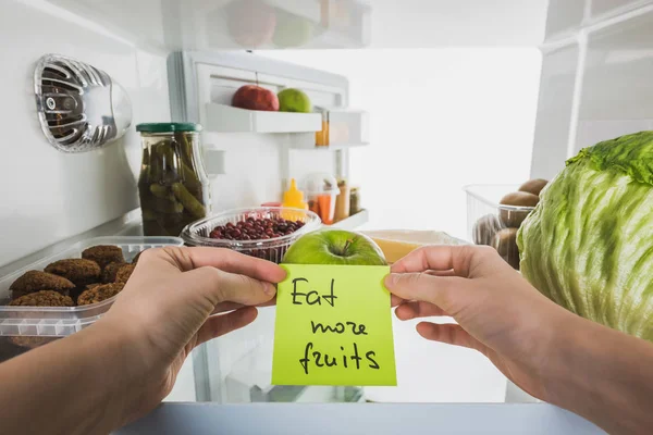 Vista cortada do cartão de exploração da mulher com comer mais frutas lettering com alimentos na geladeira isolado no branco — Fotografia de Stock